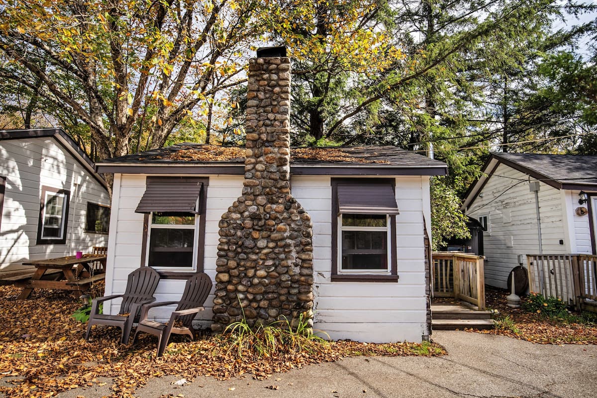Una pequeña casa con una chimenea de piedra, una terraza de madera y una silla afuera.