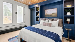 A bedroom with a large bed, a blue accent wall, built-in shelving with decorative items, and a window with blinds. - Pemi Cabins (Lincoln)