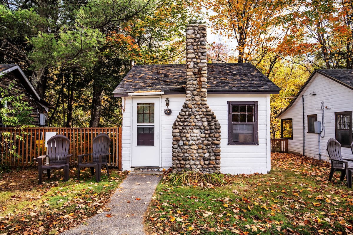 Una cabaña blanca con chimenea de piedra, rodeada de hojas otoñales, y dos sillas Adirondack en el exterior.
