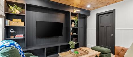 Una sala de estar moderna con televisor de pantalla plana, paneles de madera oscura, sofás verde y naranja, y una mesa de centro de madera.