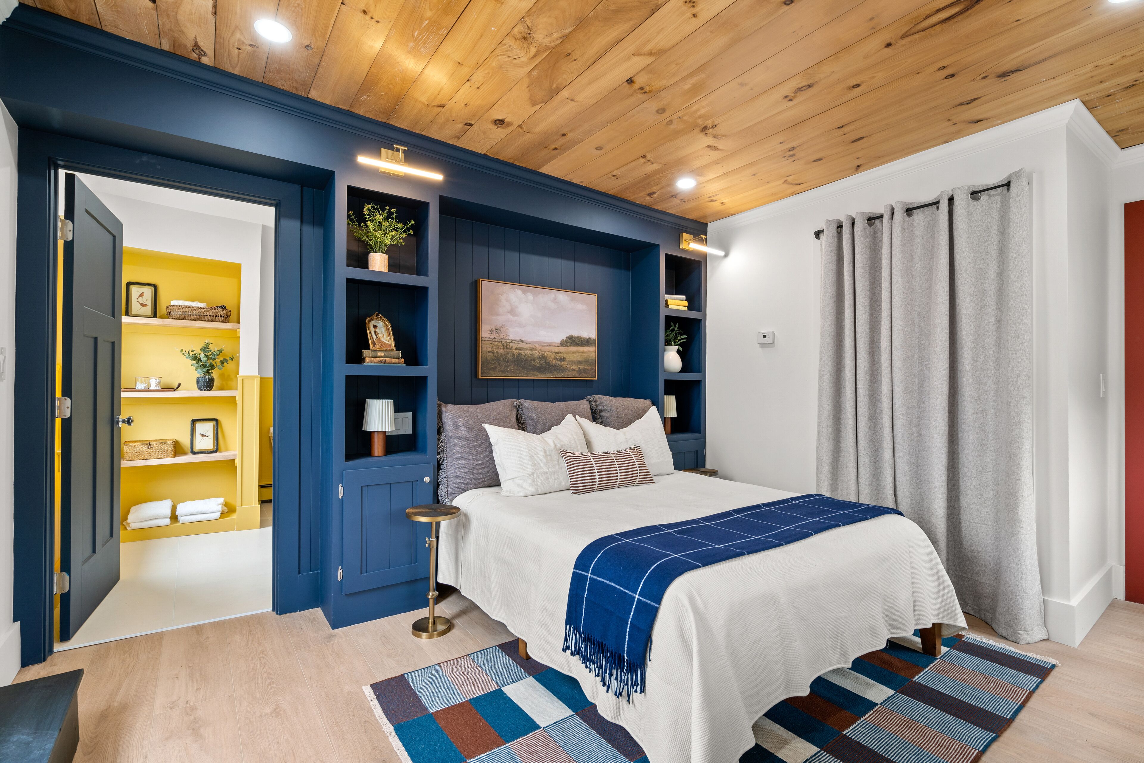 A bedroom with a wooden ceiling, a bed with a blue blanket, a nightstand, and a shelf with decorative items.