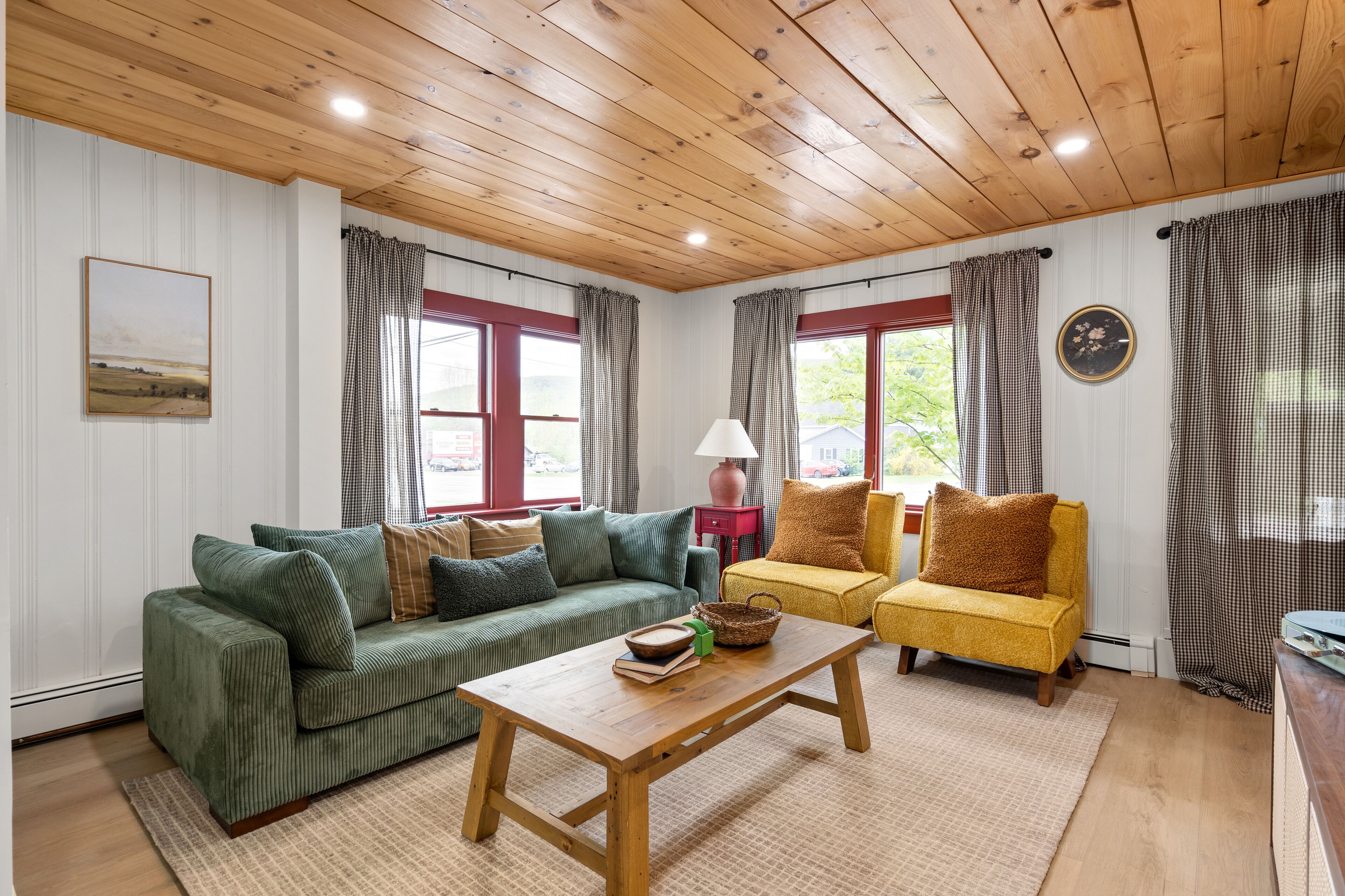 Un salon chaleureux avec un plafond en bois, des canapés verts et jaunes, une table basse en bois et une vue sur l’extérieur grâce à de grandes fenêtres.