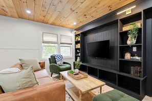 A living room with a wooden ceiling, a black entertainment unit, a green armchair, a brown leather sofa, and a flat-screen TV. - Pemi Cabins (Lincoln)