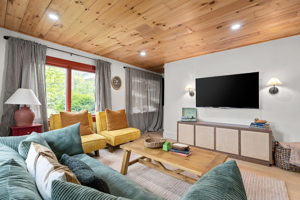 Una sala de estar acogedora con techo de madera, un conjunto de sofá, una mesa de centro, un televisor y una ventana con cortinas.