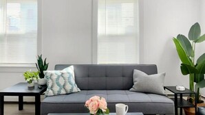 Een moderne woonkamer met een grijze bank, twee bijzettafels met planten en een groot raam met jaloezieën.