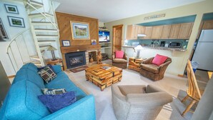 Een woonkamer met een blauwe bank, een houten salontafel, een open haard, een televisie en een keukenhoek met houten kastjes en een magnetron.