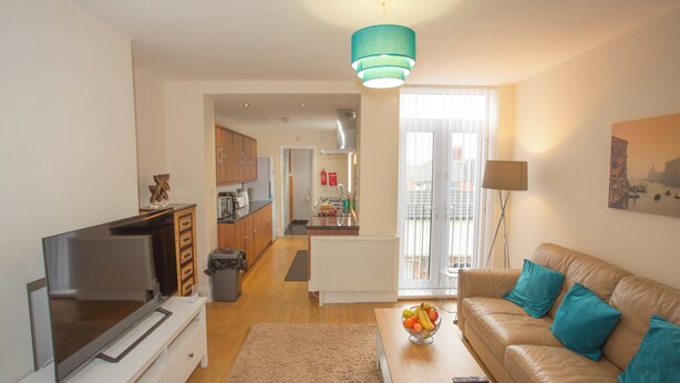 A modern living room with a TV, sofa, coffee table, and a kitchen in the background. - Norton Serviced Apartments (Stockton-on-Tees)