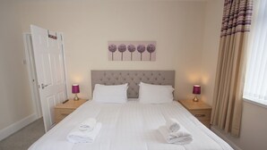 A neatly made bed with white linens and towels, a tufted headboard, two bedside tables with lamps, and a wall-mounted artwork of purple flowers. - Norton Serviced Apartments (Stockton-on-Tees)