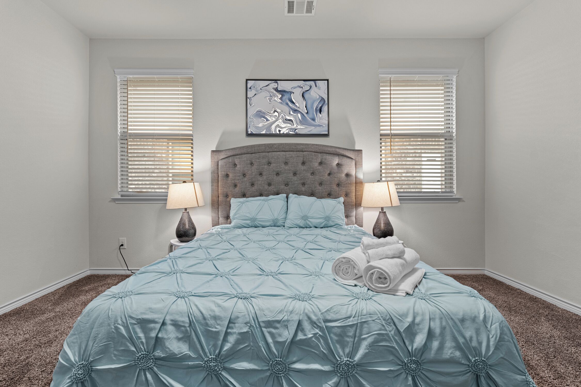 A bedroom with a large bed, two bedside lamps, a headboard, and a painting above the bed.