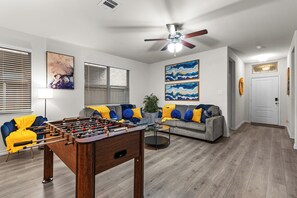 A living room with a foosball table, a gray sofa with yellow and blue pillows, a ceiling fan, and artwork on the walls. - 3BR Aubrey | Crews • Insurance • Relocation (Krugerville)