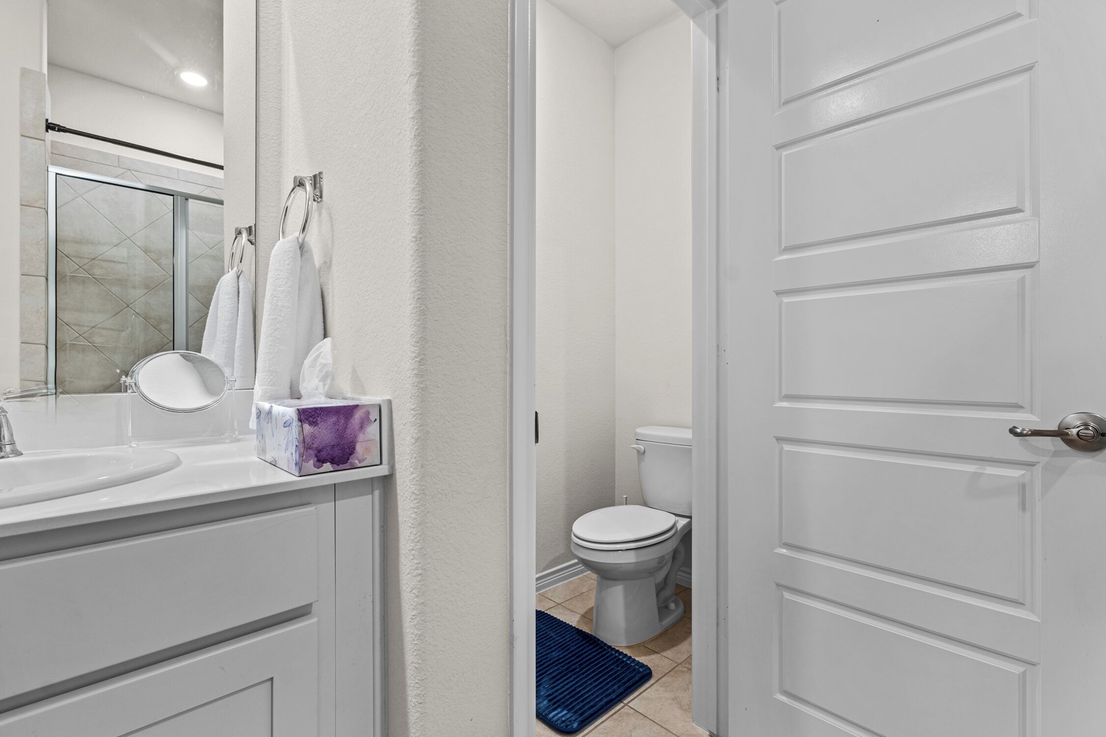 Une salle de bain avec un lavabo blanc, un miroir et des toilettes. Des serviettes sont suspendues sur un porte-serviettes et un tapis bleu est posé sur le sol.