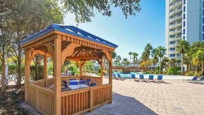 A wooden gazebo with seating, a pool area with lounge chairs, and a tall building in the background.