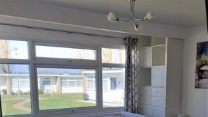 Un salon moderne avec une table à manger en verre, un canapé gris et un fauteuil en cuir noir. De grandes fenêtres offrent une vue sur un immeuble et une pelouse.
