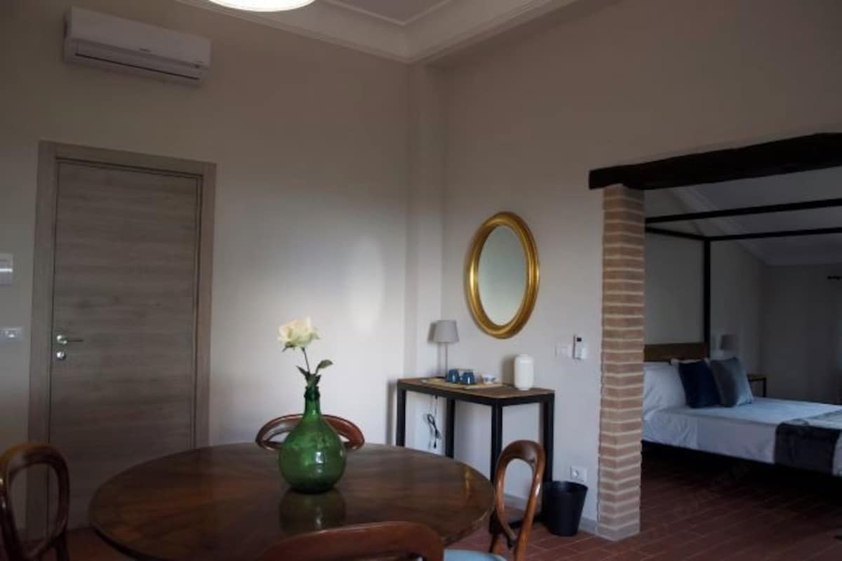 A room with a dining table, chairs, a vase with flowers, a mirror, a small table with a lamp, and a bed with pillows.