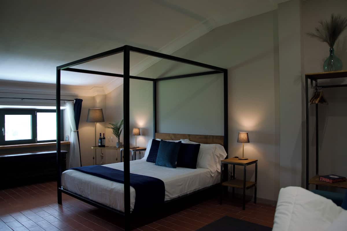 A four-poster bed with a wooden headboard, two bedside tables with lamps, a window with curtains, and a small plant on the nightstand.
