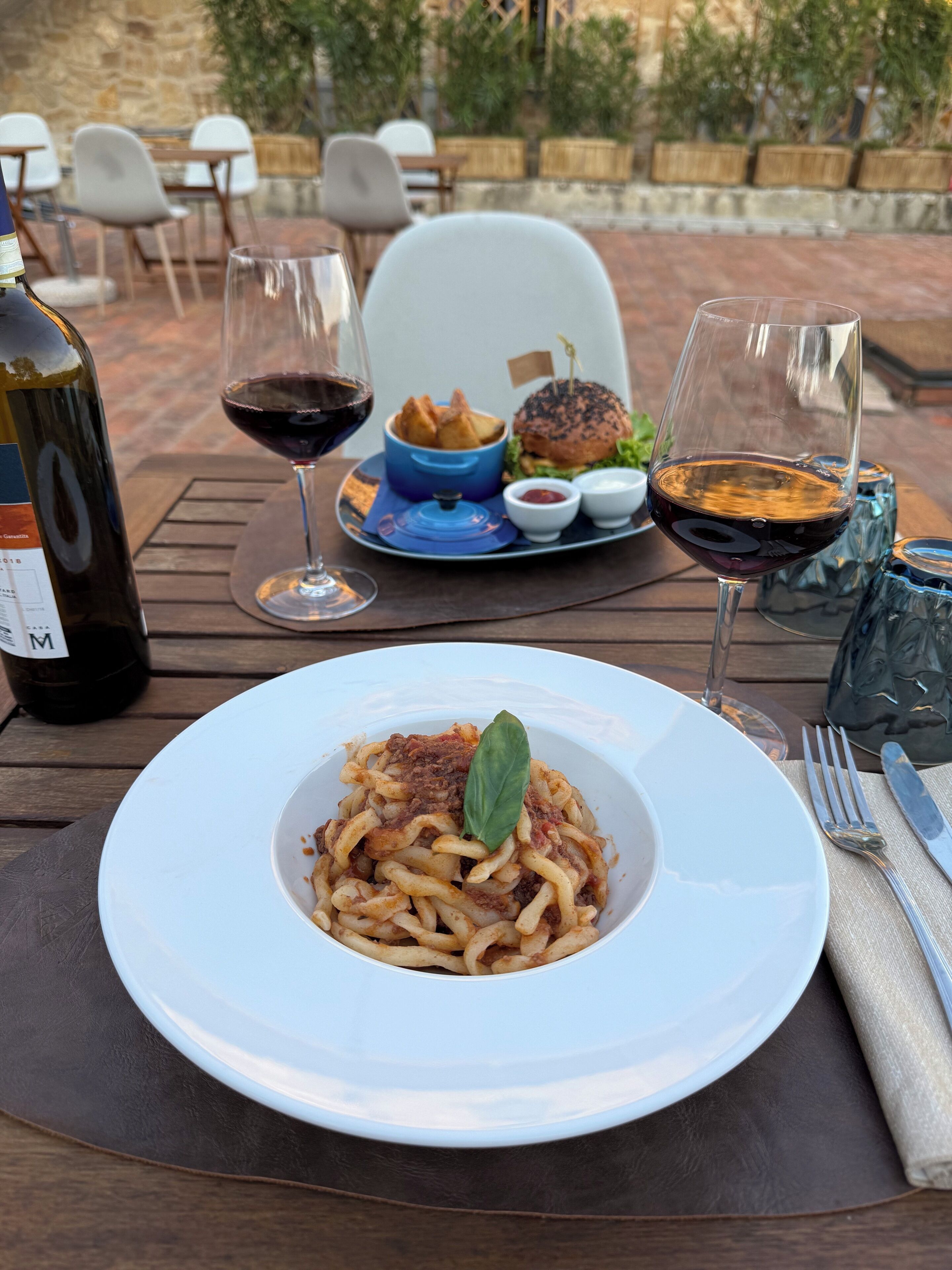 Une table à manger avec une assiette de spaghettis, un hamburger et deux verres de vin rouge.