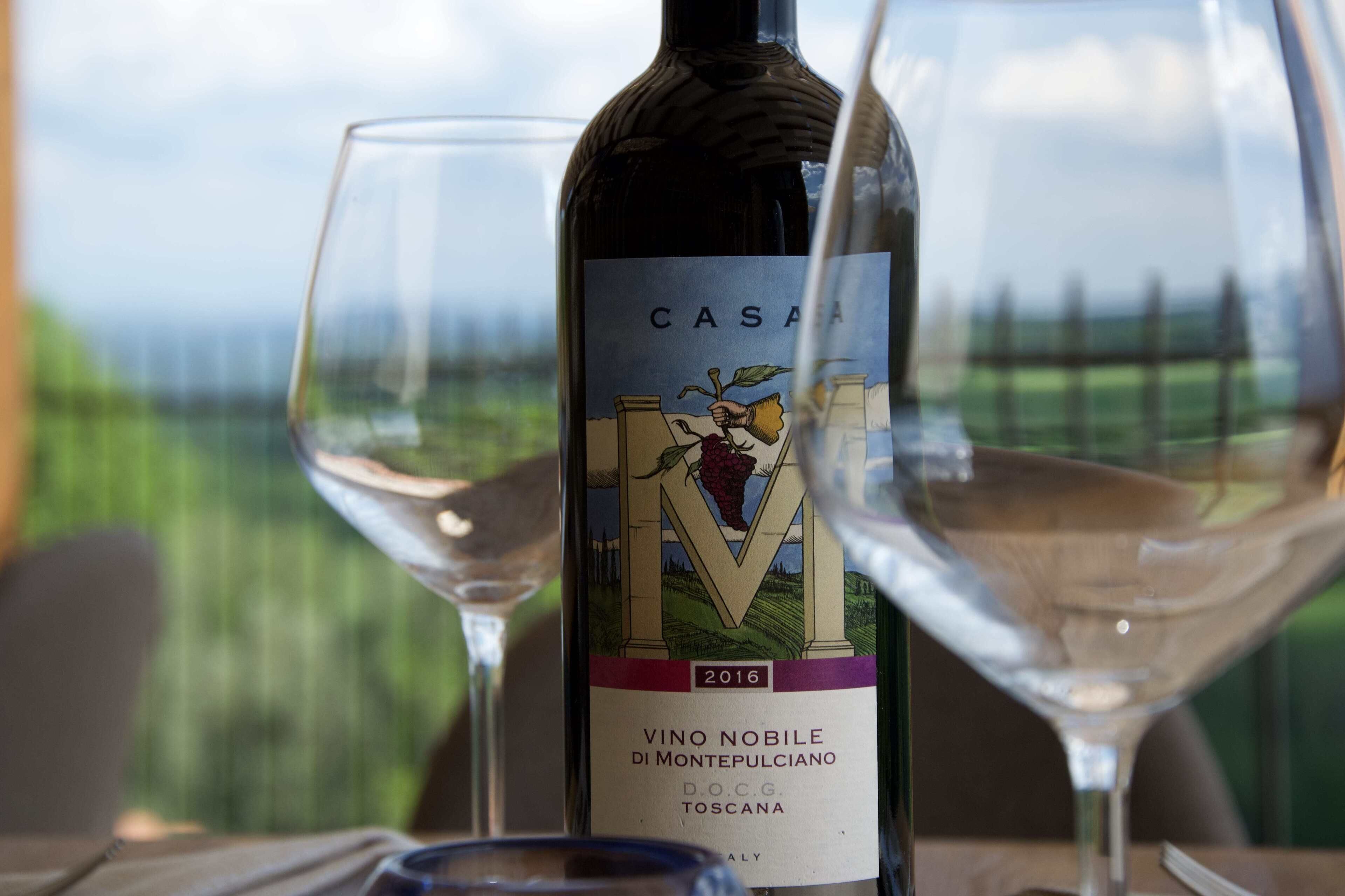 Une bouteille de vin portant l’étiquette « CASA VINO NOBILE DI MONTEPULCIANO 2016 », accompagnée de deux verres à vin, est posée sur une table.