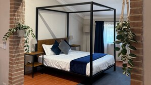 Una cama con dosel de madera, un colchón blanco y ropa de cama azul. Hay una mesita de noche pequeña con una lámpara, una planta de macramé colgante y una pared de ladrillo.