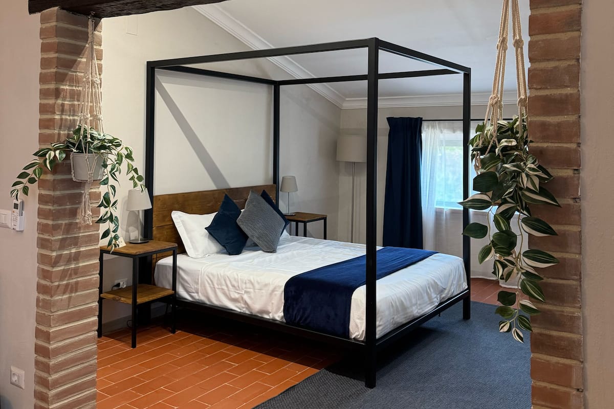 A four-poster bed with a wooden frame, a white mattress, and blue bedding. There is a small bedside table with a lamp, a hanging macrame plant, and a brick wall.