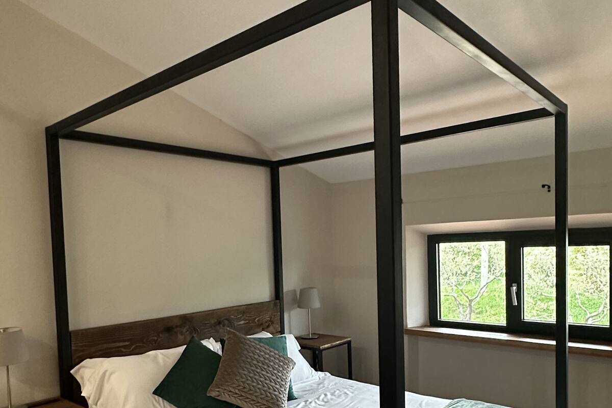 A four-poster bed with white linens and green pillows, a bedside table with a lamp, and a window with a view of greenery.