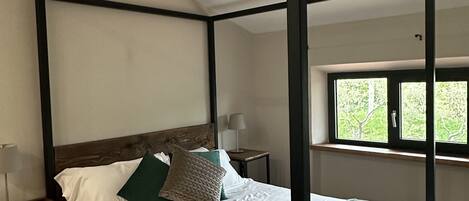 Una cama con dosel con sábanas blancas y almohadas verdes, una mesita de noche con lámpara y una ventana con vistas a la vegetación.