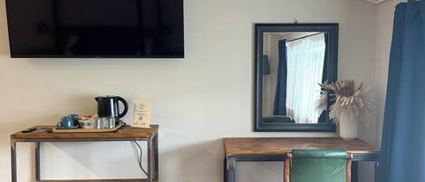 A room with a flat-screen TV mounted on the wall, a wooden desk with a chair, a small trash bin, and a tiled floor.
