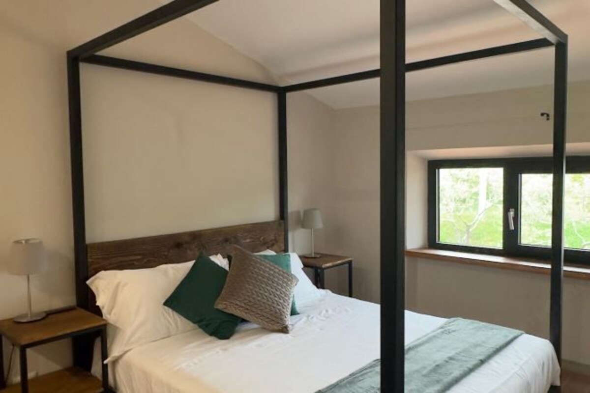 A bedroom with a four-poster bed, a window with a view of greenery, and bedside lamps.