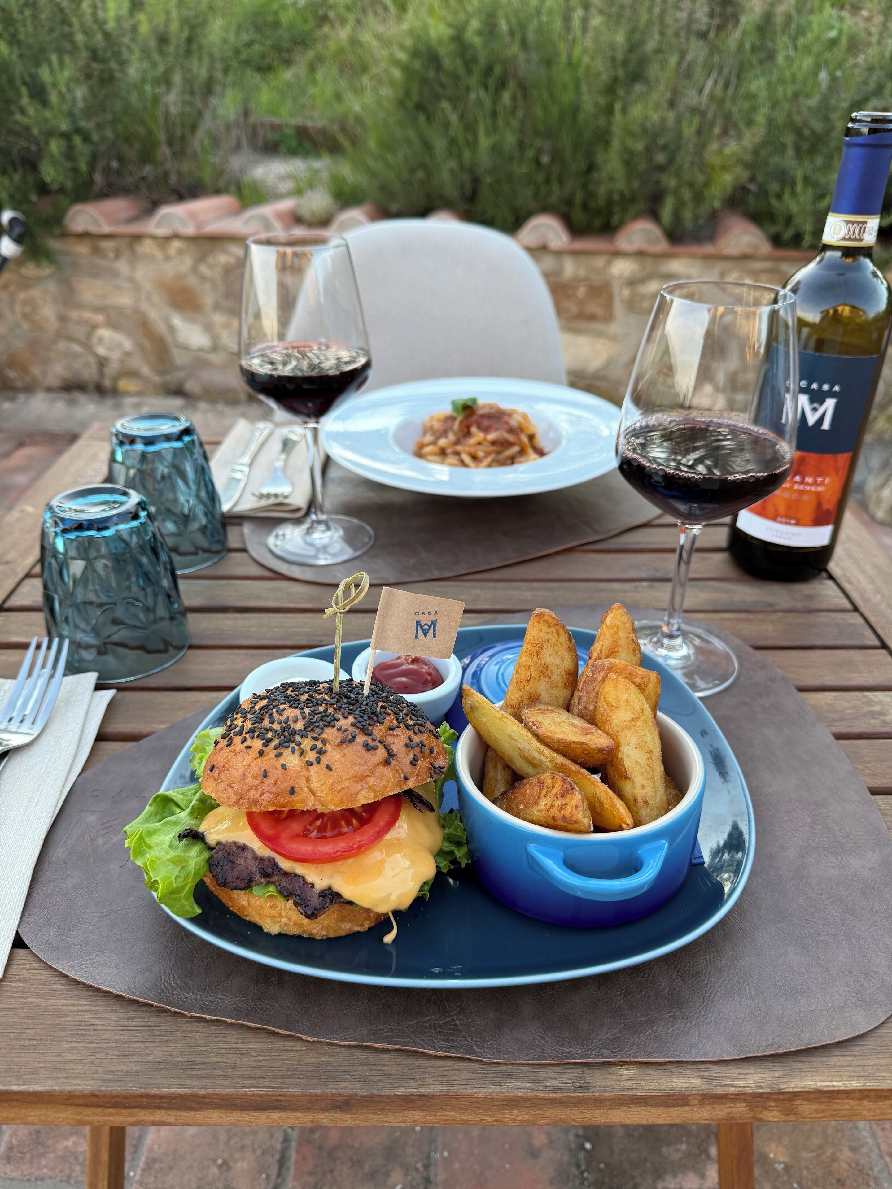 Een maaltijd op een buitentafel, bestaande uit een hamburger, friet en een glas rode wijn.