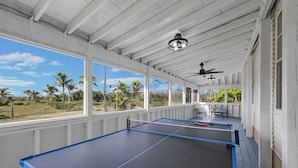 A ping pong table with a paddle, a ceiling fan, and a view of palm trees.