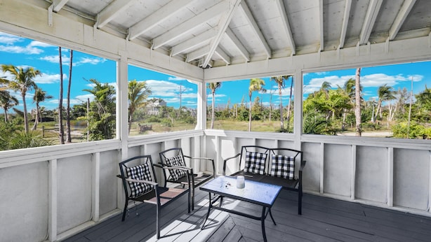 A covered outdoor seating area with a table and chairs, large windows, and a view of palm trees and greenery.