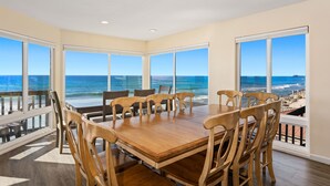 A dining area with a large table and chairs, large windows with ocean views, and a balcony with a railing. - Oceanfront Family Getaway | BBQ | Outdoor Seating (Oceanside)