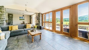 A spacious living room with a stone wall, a grey sofa, a wooden coffee table, and large windows offering a view of the countryside.