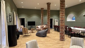 Een ruime kamer met hoge plafonds, zichtbare bakstenen kolommen, een grote flatscreen-tv, comfortabele zithoek met banken en stoelen, en een lange eettafel.