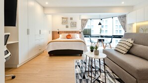 A modern bedroom with a bed, a sofa, a small table, and a balcony with a view of buildings. - ALU Apartments - Miraflores Central (Lima)