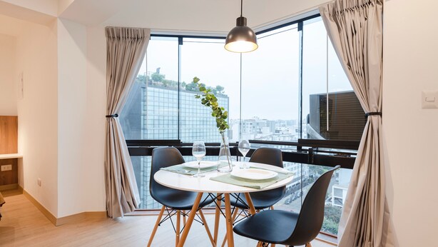 A dining area with a round table, black chairs, a hanging pendant light, and a view of the city through large windows. - ALU Apartments - Miraflores Central (Lima)