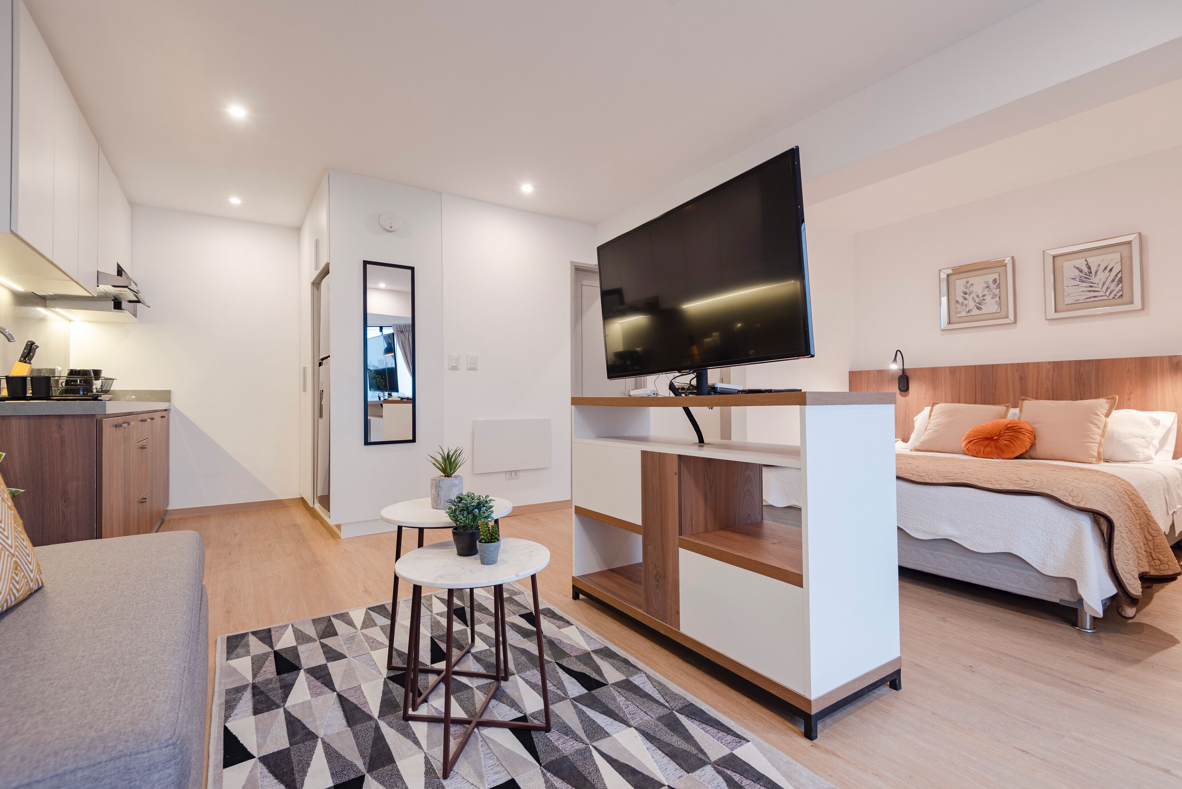 A modern hotel room with a bed, a TV, a small kitchenette, and a seating area with a rug.