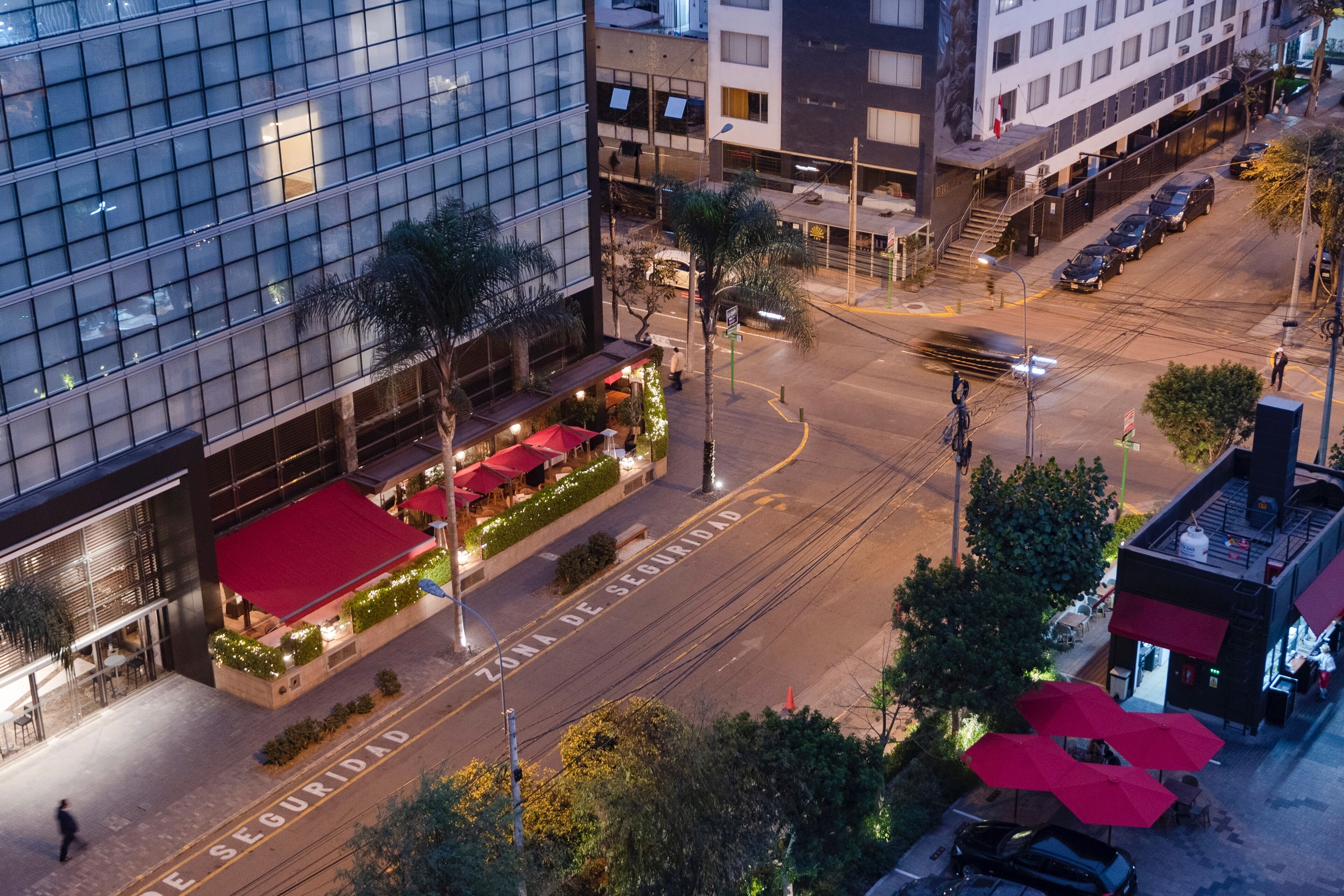 A multi-story building with a glass facade, outdoor seating area with red umbrellas, and a security zone marking on the street.