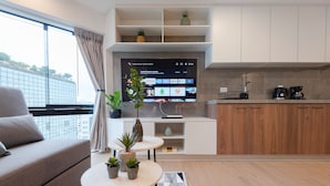 A modern living room with a sofa, a coffee table with plants, a TV displaying a streaming service interface, and a kitchen area with wooden cabinets and a coffee machine. - ALU Apartments - Miraflores Central (Lima)