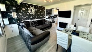 A modern living room with a sofa, a dining table with chairs, a kitchen area, and a decorative wall clock.