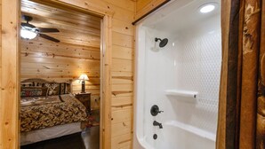 Et interiør af en træhytte med en seng, en ventilator i loftet, et natbord, en lampe og et hvidt badekar.