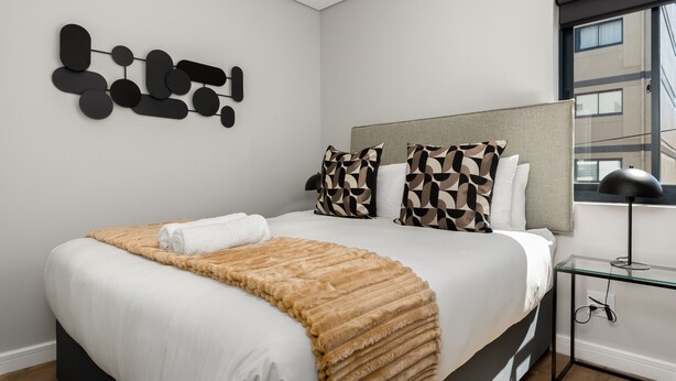 Un dormitorio moderno con una cama grande, almohadones estampados, una obra de arte en la pared y una mesita de noche con una lámpara.