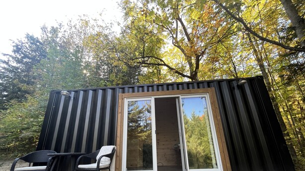 Une cabane compacte et moderne, dotée d’une porte-fenêtre coulissante, d’un petit patio avec une chaise et d’une terrasse en bois.