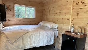 Une pièce de cabane en bois comprenant un lit, un petit réfrigérateur, une fenêtre et un sac posé sur le sol.