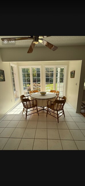 Un coin repas avec une table ronde, quatre chaises, un ventilateur de plafond et de grandes fenêtres.