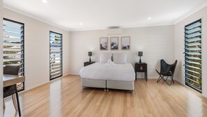 A modern bedroom with a large bed, two bedside tables, a chair, and a view through a window with blinds. - 22 Seaview Terrace Moffat Beach (Moffat Beach)