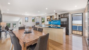 A modern kitchen with a central island, dining area with a wooden table and chairs, and a view of the outside through a sliding door. - 22 Seaview Terrace Moffat Beach (Moffat Beach)