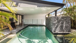 A modern outdoor pool area with a clear pool, a stone wall, and a covered seating area with a table and chairs. - 22 Seaview Terrace Moffat Beach (Moffat Beach)