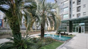 A modern building with a glass facade, a courtyard with a fountain, and palm trees.