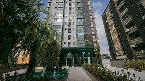 A modern high-rise building with a central entrance, surrounded by greenery and a small water feature.