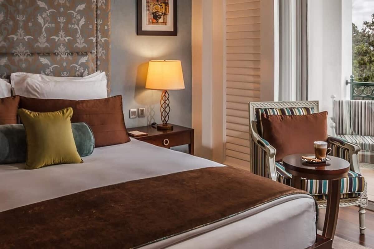 A hotel room with a large bed, a nightstand with a lamp, a small table with a cup, and a chair with striped cushions.